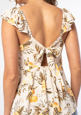 [Color: Natural/Yellow] A woman wearing a natural and yellow floral fruit print bohemian top with short flutter sleeves and a tie back detail. 