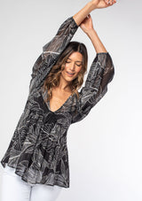 [Color: Black/Ivory] A model wearing a black leaf print sheer chiffon bohemian blouse with gold metallic clip dot details throughout. With long voluminous sleeves and a ruffle trimmed waist.