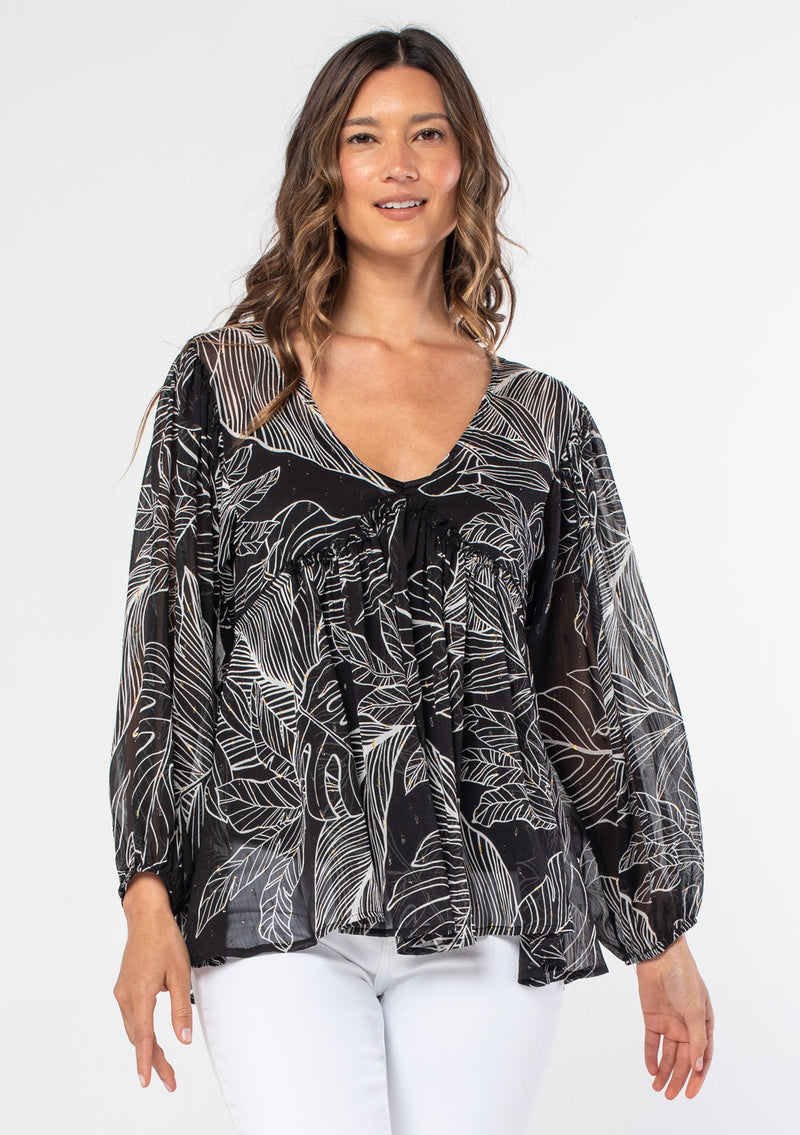 [Color: Black/Ivory] A model wearing a black leaf print sheer chiffon bohemian blouse with gold metallic clip dot details throughout. With long voluminous sleeves and a ruffle trimmed waist.