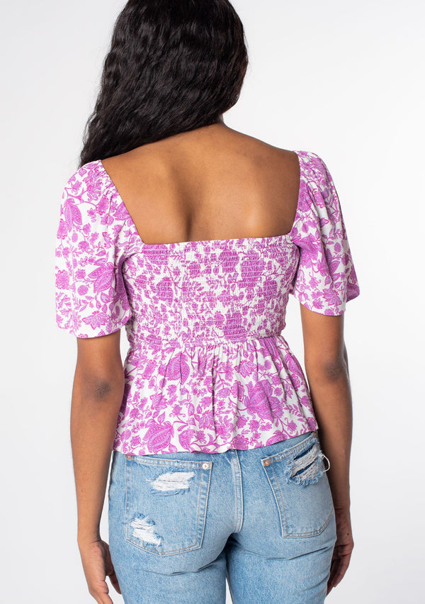 [Color: Ivory/Lilac] A model wearing a purple and white floral print bohemian peplum top with a slim fit smocked bodice and short flutter sleeves. 