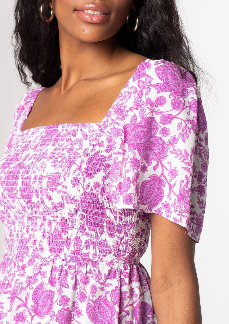[Color: Ivory/Lilac] A model wearing a purple and white floral print bohemian peplum top with a slim fit smocked bodice and short flutter sleeves. 
