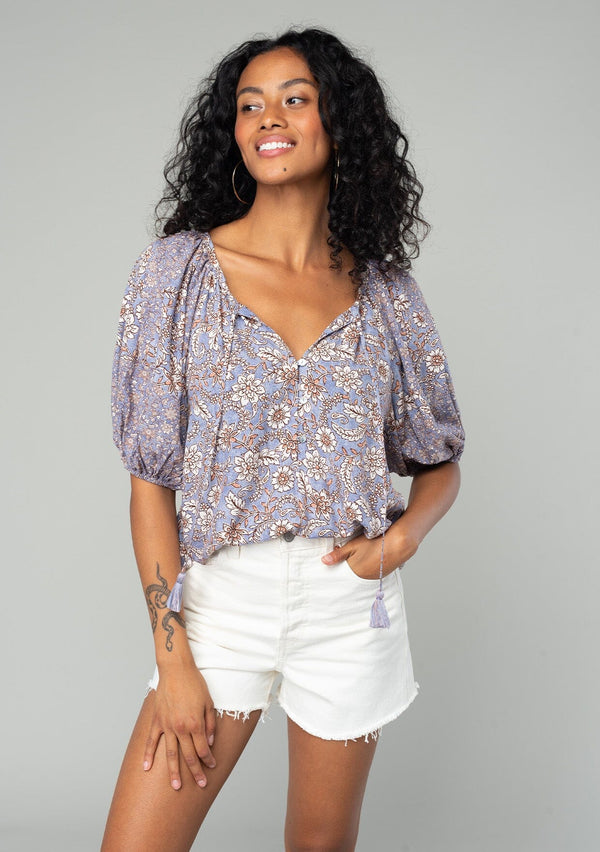 [Color: Grey/Natural] A front facing image of a brunette model wearing a classic bohemian blouse in a grey and purple mixed floral print. With short puff sleeves, a loop button front, and a split v neckline with tassel ties. 