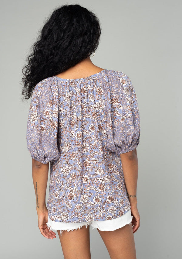 [Color: Grey/Natural] A back facing image of a brunette model wearing a classic bohemian blouse in a grey and purple mixed floral print. With short puff sleeves, a loop button front, and a split v neckline with tassel ties. 
