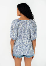 [Color: Ivory/Blue] A back facing image of a brunette model wearing a bohemian spring blouse with short puff sleeves, a button front, a split v neckline, and tassel ties. 