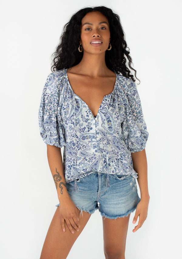 [Color: Ivory/Blue] A half body front facing image of a brunette model wearing a bohemian spring blouse with short puff sleeves, a button front, a split v neckline, and tassel ties. 