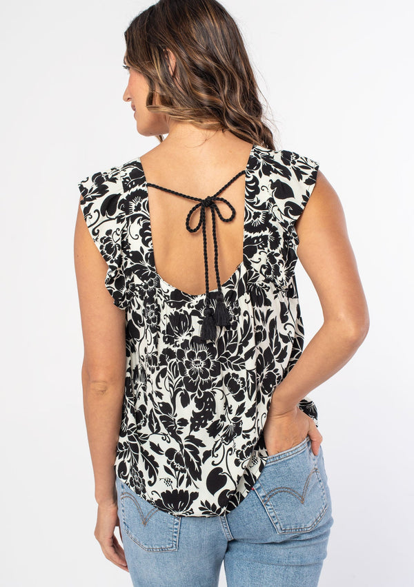 [Color: Ivory/Black] A woman wearing a white and black tropical floral print flowy bohemian top with short flutter sleeves and a square neckline. 