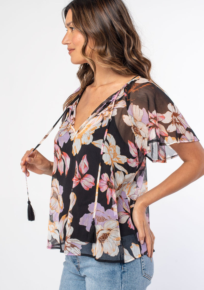 [Color: Black/Lilac] A woman wearing a sheer black bohemian top with a purple floral print. With short flutter sleeves and a split v neckline with tassel ties. 