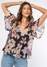 [Color: Black/Lilac] A woman wearing a sheer black bohemian top with a purple floral print. With short flutter sleeves and a split v neckline with tassel ties. 