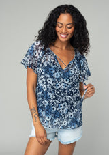 [Color: Indigo/Blue] A half body front facing image of a brunette model wearing a bohemian sheer chiffon top in a blue floral print. With short flutter sleeves, a split neckline with tassel ties, and a flowy relaxed fit. 