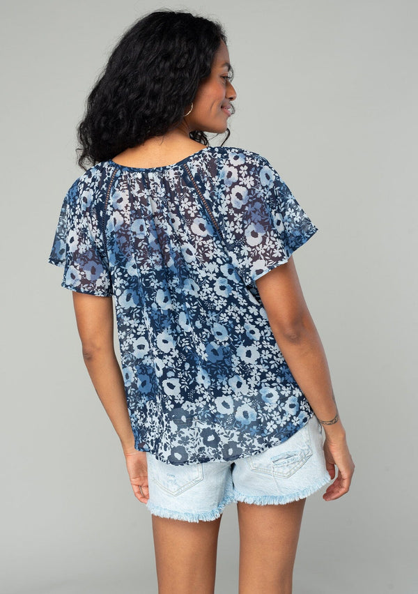 [Color: Indigo/Blue] A back facing image of a brunette model wearing a bohemian sheer chiffon top in a blue floral print. With short flutter sleeves, a split neckline with tassel ties, and a flowy relaxed fit. 