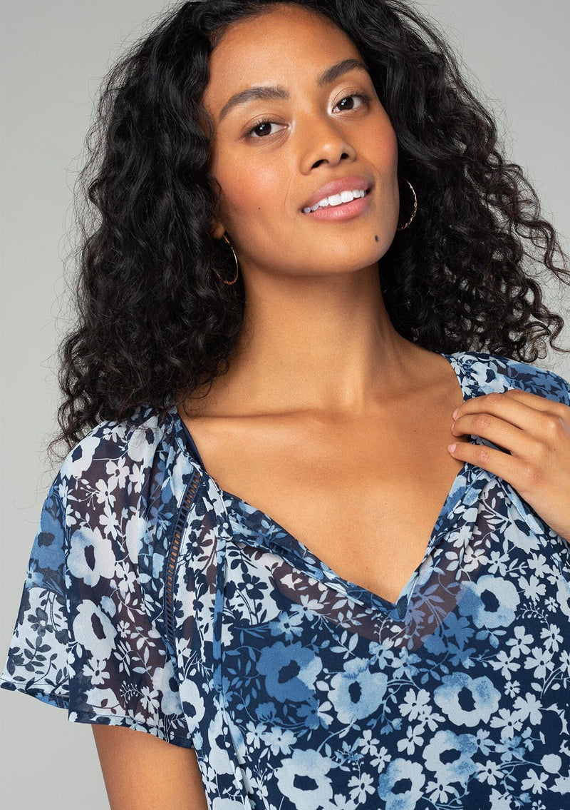 [Color: Indigo/Blue] A close up front facing image of a brunette model wearing a bohemian sheer chiffon top in a blue floral print. With short flutter sleeves, a split neckline with tassel ties, and a flowy relaxed fit. 