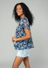 [Color: Indigo/Blue] A side facing image of a brunette model wearing a bohemian sheer chiffon top in a blue floral print. With short flutter sleeves, a split neckline with tassel ties, and a flowy relaxed fit. 