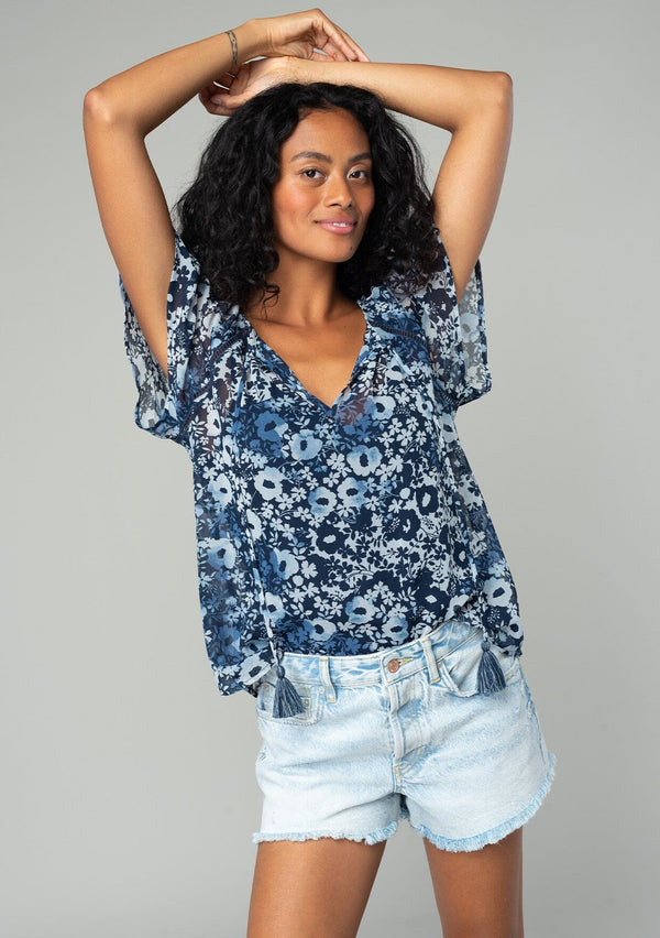 [Color: Indigo/Blue] A front facing image of a brunette model wearing a bohemian sheer chiffon top in a blue floral print. With short flutter sleeves, a split neckline with tassel ties, and a flowy relaxed fit. 