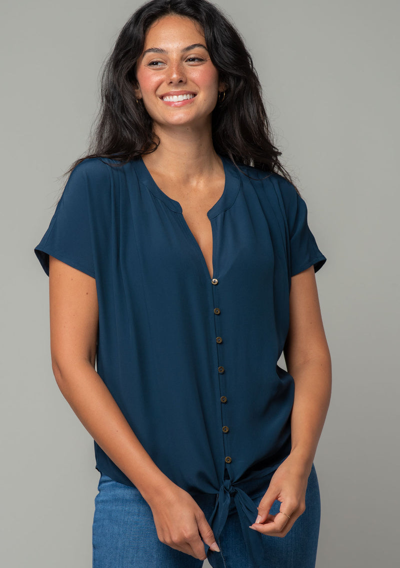 [Color: Midnight] A front facing image of a brunette model wearing a soft and silky navy blue short sleeve top with a button front and tie waist detail. 