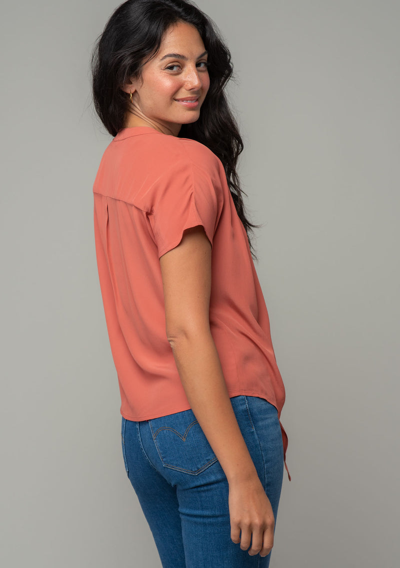 [Color: Rosewood] A side facing image of a brunette model wearing a soft and silky dusty rose pink short sleeve top with a button front and tie waist detail. The model is looking over her shoulder. 
