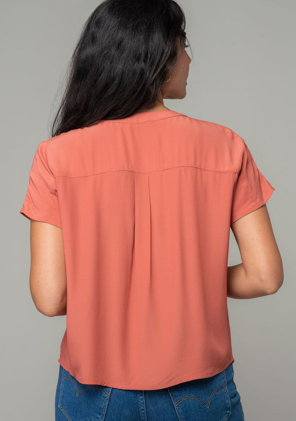 [Color: Rosewood] A back facing image of a brunette model wearing a soft and silky dusty rose pink short sleeve top with a button front and tie waist detail. 