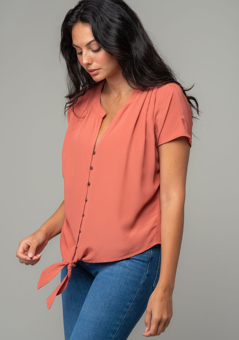 [Color: Rosewood] A side facing image of a brunette model wearing a soft and silky dusty rose pink short sleeve top with a button front and tie waist detail. 