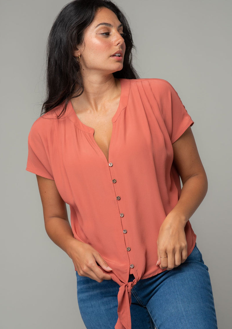 [Color: Rosewood] A front facing image of a brunette model wearing a soft and silky dusty rose pink short sleeve top with a button front and tie waist detail. 