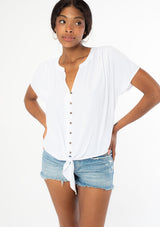 [Color: Chalk] A front facing image of a black model wearing a soft and silky white short sleeve top with a button front and tie waist detail. The model has her hands on her hips.