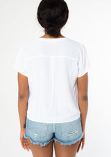 [Color: Chalk] A back facing image of a black model wearing a soft and silky white short sleeve top with a button front and tie waist detail. 