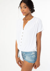 [Color: Chalk] A side facing image of a black model wearing a soft and silky white short sleeve top with a button front and tie waist detail. 