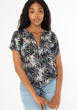 [Color: Black/Blue] A front facing image of a black model wearing a multicolor black and blue palm print short sleeve top with a button front and a tie front waist detail. 