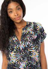 [Color: Black/Blue] A close up front facing image of a black model wearing a multicolor black and blue palm print short sleeve top with a button front and a tie front waist detail. 