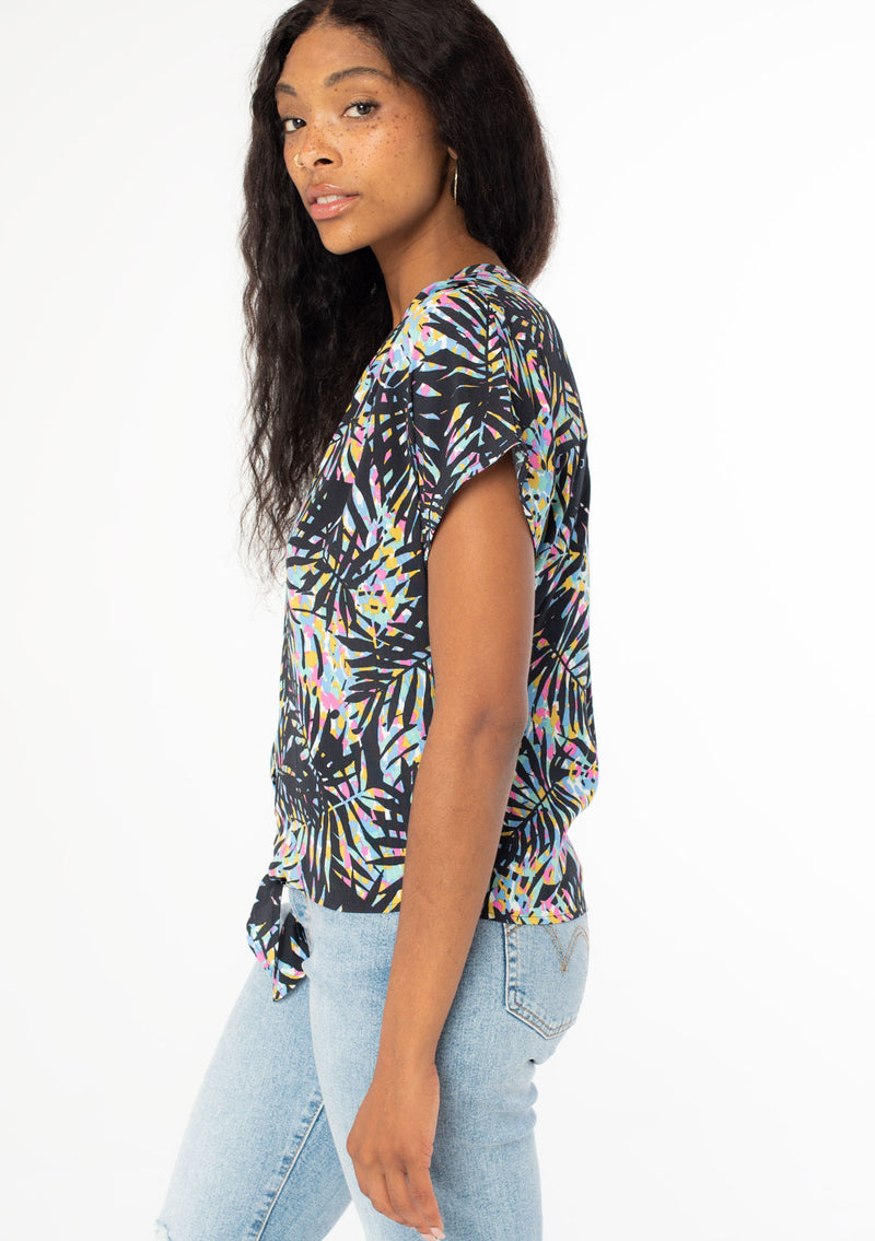 [Color: Black/Blue] A side facing image of a black model wearing a multicolor black and blue palm print short sleeve top with a button front and a tie front waist detail. 