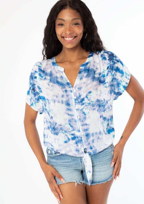 [Color: Navy/Teal] A front facing image of a black model wearing a blue and white watercolor floral print short sleeve top with a button front and a tie front detail at the waist. 