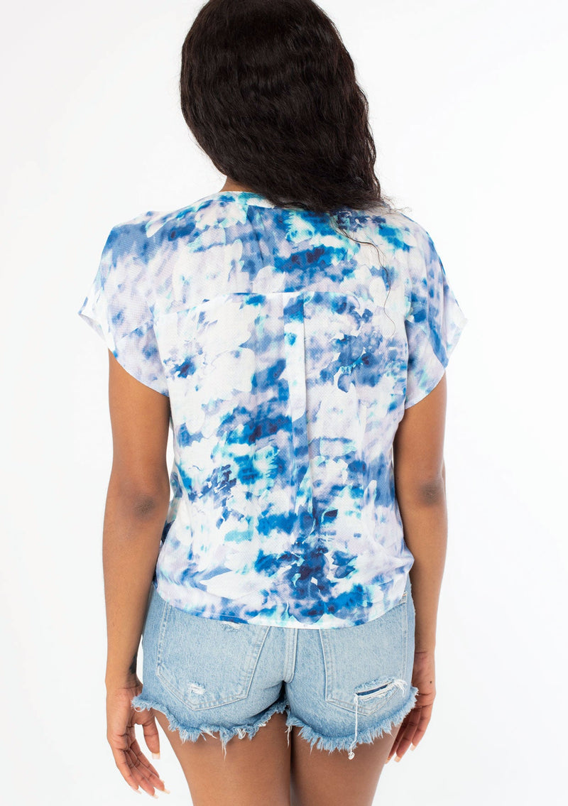 [Color: Navy/Teal] A back facing image of a black model wearing a blue and white watercolor floral print short sleeve top with a button front and a tie front detail at the waist. 