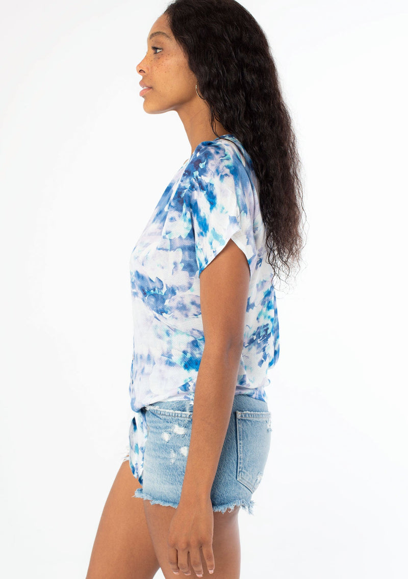 [Color: Navy/Teal] A side facing image of a black model wearing a blue and white watercolor floral print short sleeve top with a button front and a tie front detail at the waist. 