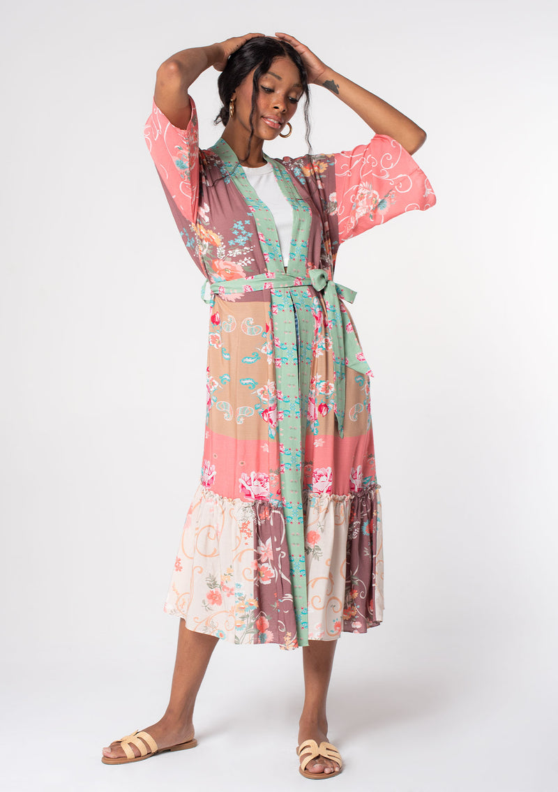 [Color: Rose/Mocha] A woman wearing a pink floral duster length robe with half length kimono sleeves, a self tie waist belt, and a ruffle trimmed tiered hemline. 