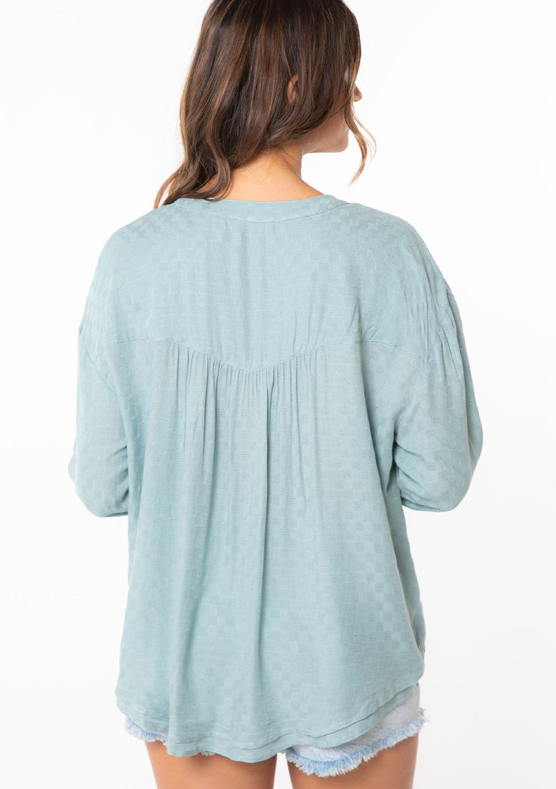 [Color: Dusty Teal] A model wearing a classic lightweight textured teal checkered jacquard long rolled tab sleeve blouse. With a notched v neckline and boxy relaxed fit.