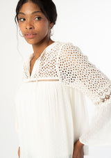 [Color: Vanilla] A close up side facing image of a black model wearing an off white sheer bohemian blouse with long bell sleeves, tassel ties, and sheer eyelet lace detail. 