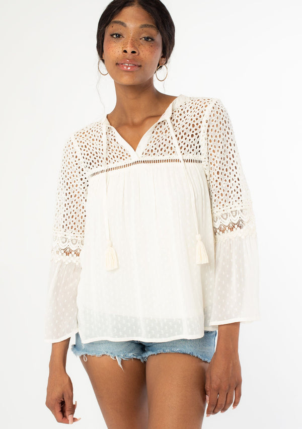 [Color: Vanilla] A front facing image of a black model wearing an off white sheer bohemian blouse with long bell sleeves, tassel ties, and sheer eyelet lace detail. 