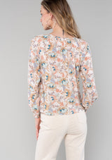 [Color: Natural/Coral] A back facing image of a blonde model wearing a bohemian blouse in a natural and coral floral print. With long sleeves, a round neckline, a self covered button front, and a tie front waist. 