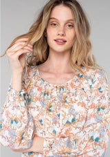 [Color: Natural/Coral] A close up front facing image of a blonde model wearing a bohemian blouse in a natural and coral floral print. With long sleeves, a round neckline, a self covered button front, and a tie front waist. 