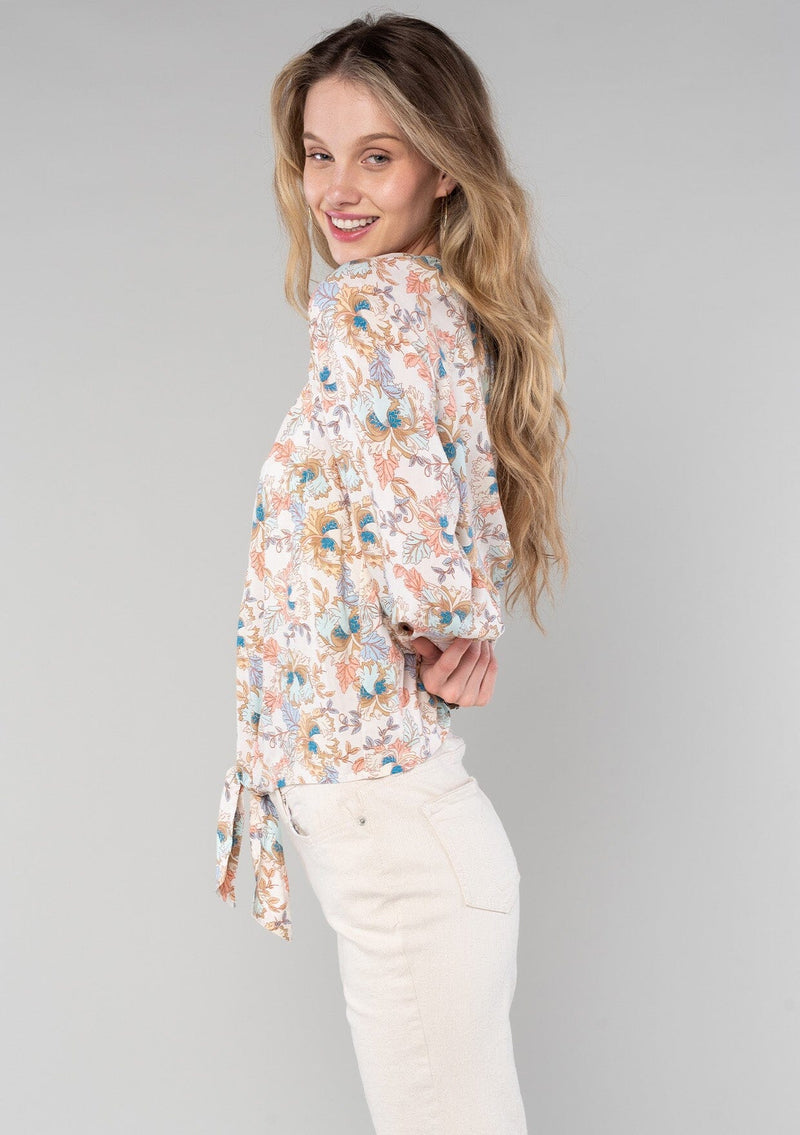 [Color: Natural/Coral] A side facing image of a blonde model wearing a bohemian blouse in a natural and coral floral print. With long sleeves, a round neckline, a self covered button front, and a tie front waist. 