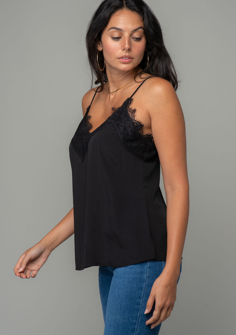 [Color: Black] A side facing image of a brunette model wearing a black lace trim camisole with a v neckline, adjustable spaghetti straps, and a flowy relaxed fit. 