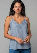 [Color: Grey] A front facing image of a brunette model wearing a light grey clip dot camisole with a lace trim v neckline and adjustable spaghetti straps. The model is wearing gold hoop earrings and a gold necklace.