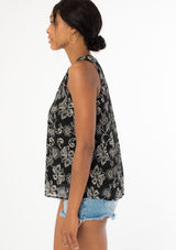 [Color: Black/Natural] A side facing image of a black model wearing a classic sleeveless bohemian tank top in a black and natural, off white floral print. A flowy chiffon top with a button front and loop button trimmed v neckline. 