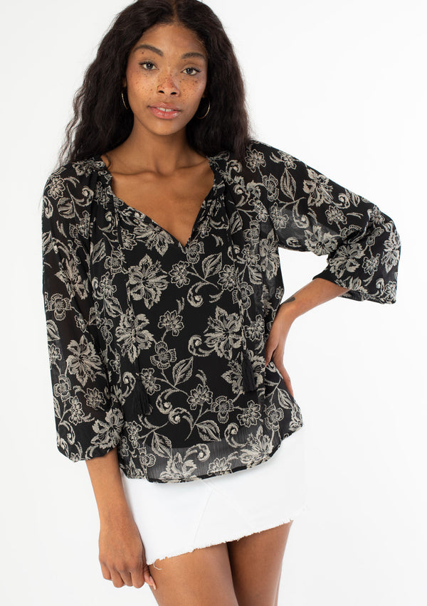 [Color: Black/Natural] A front facing image of a black model wearing a sheer chiffon bohemian blouse in a black and natural floral print. With long sleeves, a flowy silhouette, and a split v neckline with tassel neck ties. 