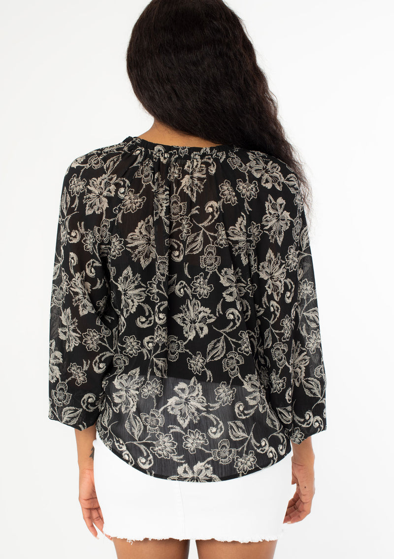 [Color: Black/Natural] A back facing image of a black model wearing a sheer chiffon bohemian blouse in a black and natural floral print. With long sleeves, a flowy silhouette, and a split v neckline with tassel neck ties. 
