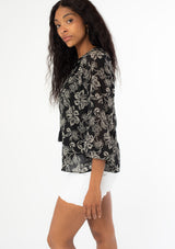 [Color: Black/Natural] A side facing image of a black model wearing a sheer chiffon bohemian blouse in a black and natural floral print. With long sleeves, a flowy silhouette, and a split v neckline with tassel neck ties. 