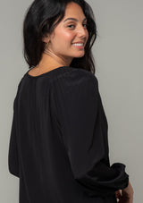 [Color: Black] A close up back facing image of a brunette model wearing a soft and silky black bohemian blouse with voluminous long sleeves, a plunging surplice v neckline, and an elastic front waist. 