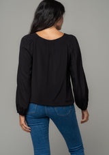 [Color: Black] A back facing image of a brunette model wearing a soft and silky black bohemian blouse with voluminous long sleeves, a plunging surplice v neckline, and an elastic front waist. 