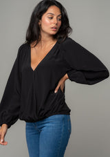 [Color: Black] A side facing image of a brunette model wearing a soft and silky black bohemian blouse with voluminous long sleeves, a plunging surplice v neckline, and an elastic front waist. 
