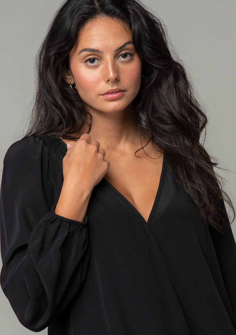 [Color: Black] A close up front facing image of a brunette model wearing a soft and silky black bohemian blouse with voluminous long sleeves, a plunging surplice v neckline, and an elastic front waist. 