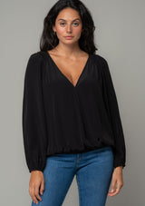 [Color: Black] A front facing image of a brunette model wearing a soft and silky black bohemian blouse with voluminous long sleeves, a plunging surplice v neckline, and an elastic front waist. 