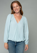 [Color: Sky] A front facing image of a red headed model wearing a soft and silky light blue bohemian blouse with voluminous long sleeves, a plunging surplice v neckline, and an elastic front waist.